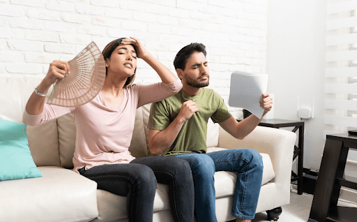 Couple fanning themselves in their living room because air conditioner is blowing warm air.