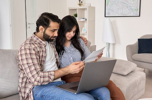 A man and woman sitting on a couch. There are looking at a laptop and sheet of paper.