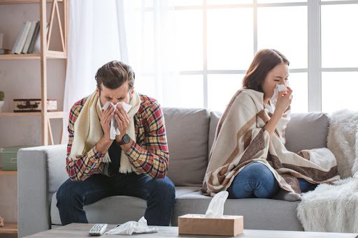 A man and woman sitting on a couch. They're both blowing their noses.