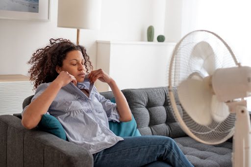 A woman adjusting the collar of her shirt as she sits in front of a fan while sitting on a couch.