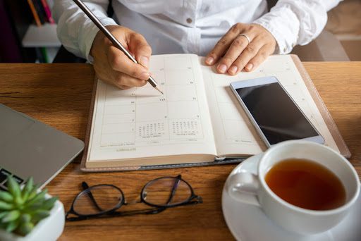 A close-up of a person's hands as they write in a planner with a phone and a cup of tea on the table.